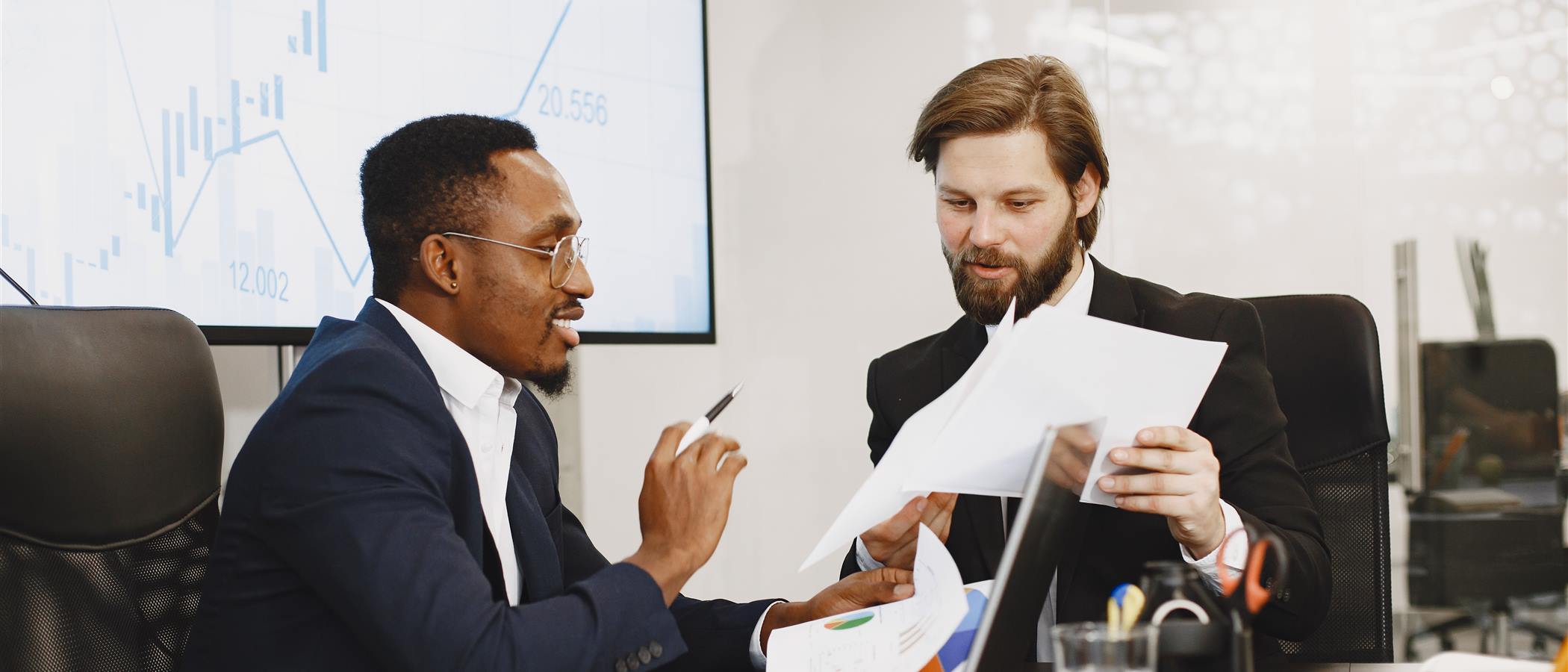 Two colleagues passing billing documents across a boardroom table in front of a revenue chart